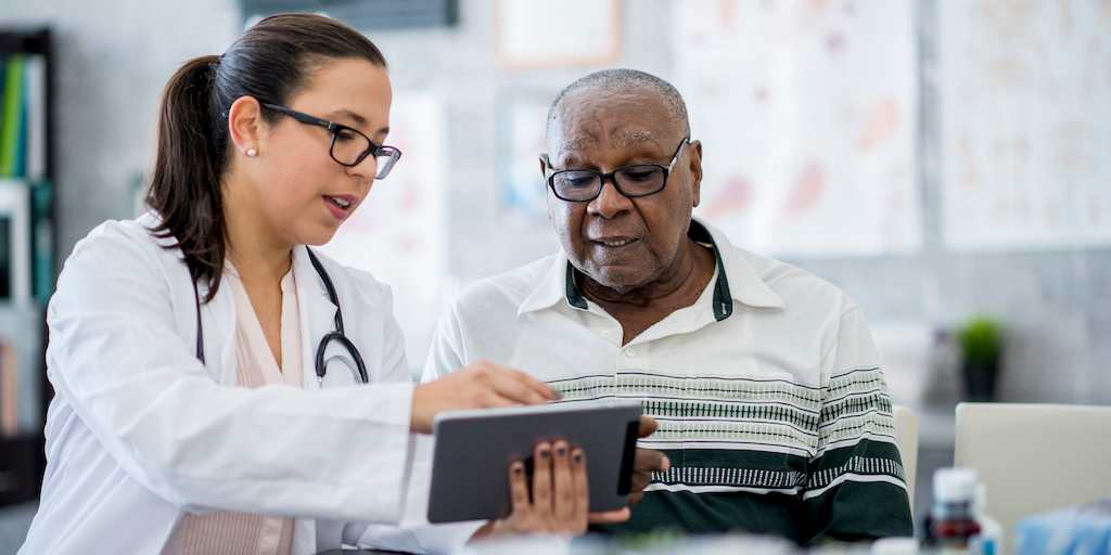 A senior man of African descent is indoors in a hospital room. He is watching his female doctor using a tablet computer. She is explaining a medication schedule to him.