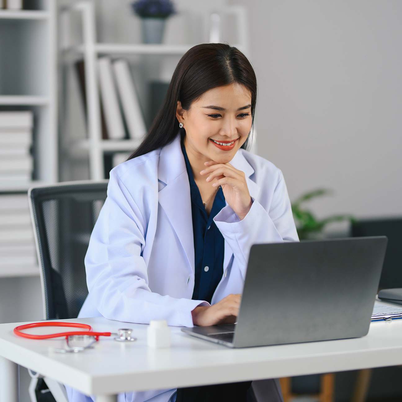 Doctor in a white lab coat is sitting at a desk with a laptop, smiling and happy.