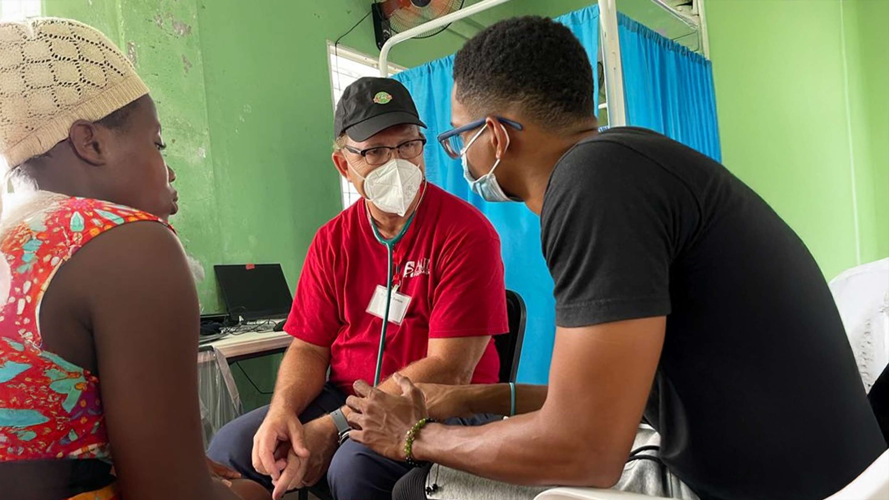 Physician wearing a mask and red shirt speaks with a patient and interpreter during a medical consultation in a community clinic.