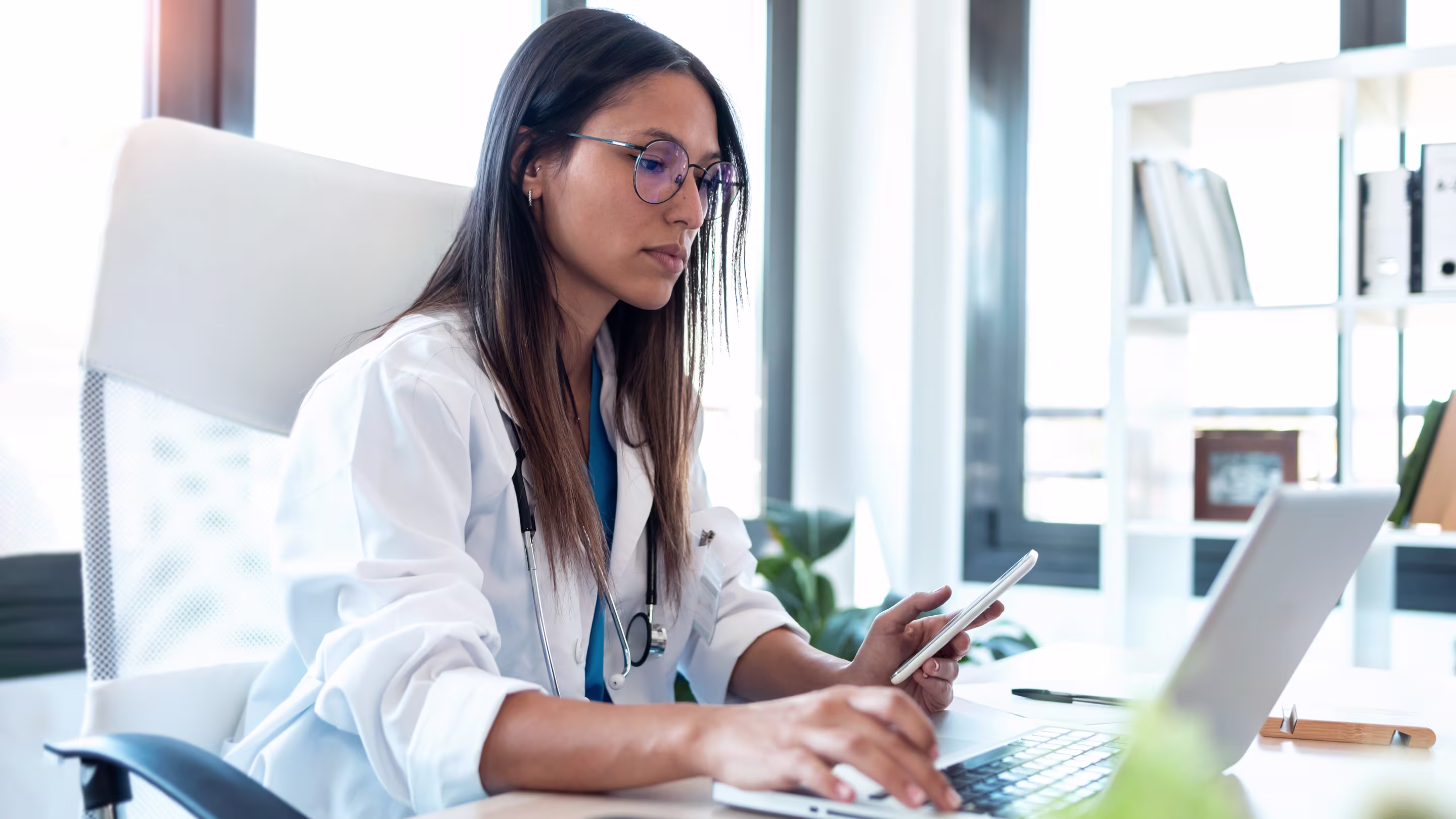 Image of confident young female doctor using her mobile phone while working on laptop in the consultation.