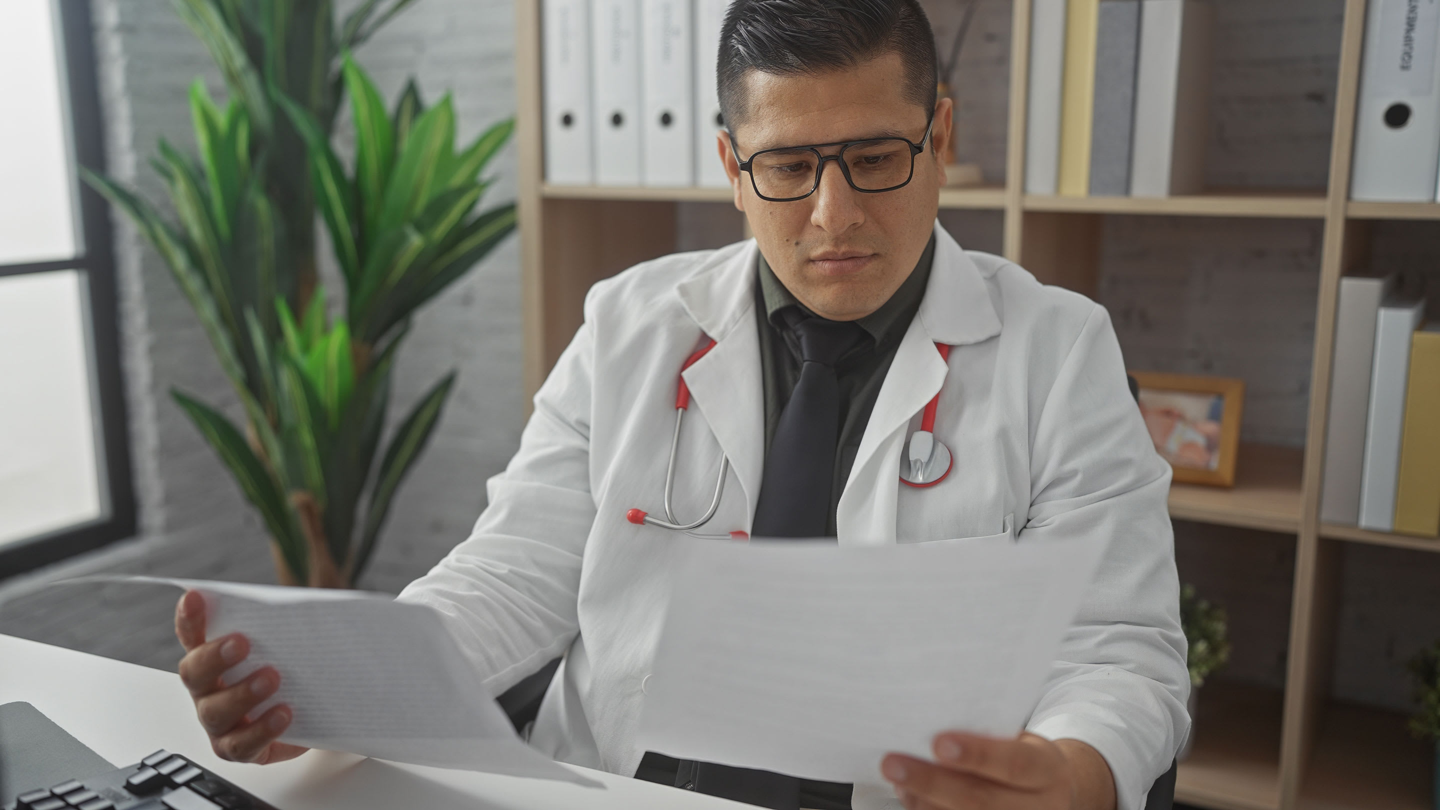Image of a focused hispanic man in a white lab coat reviews documents at his desk in a medical office.