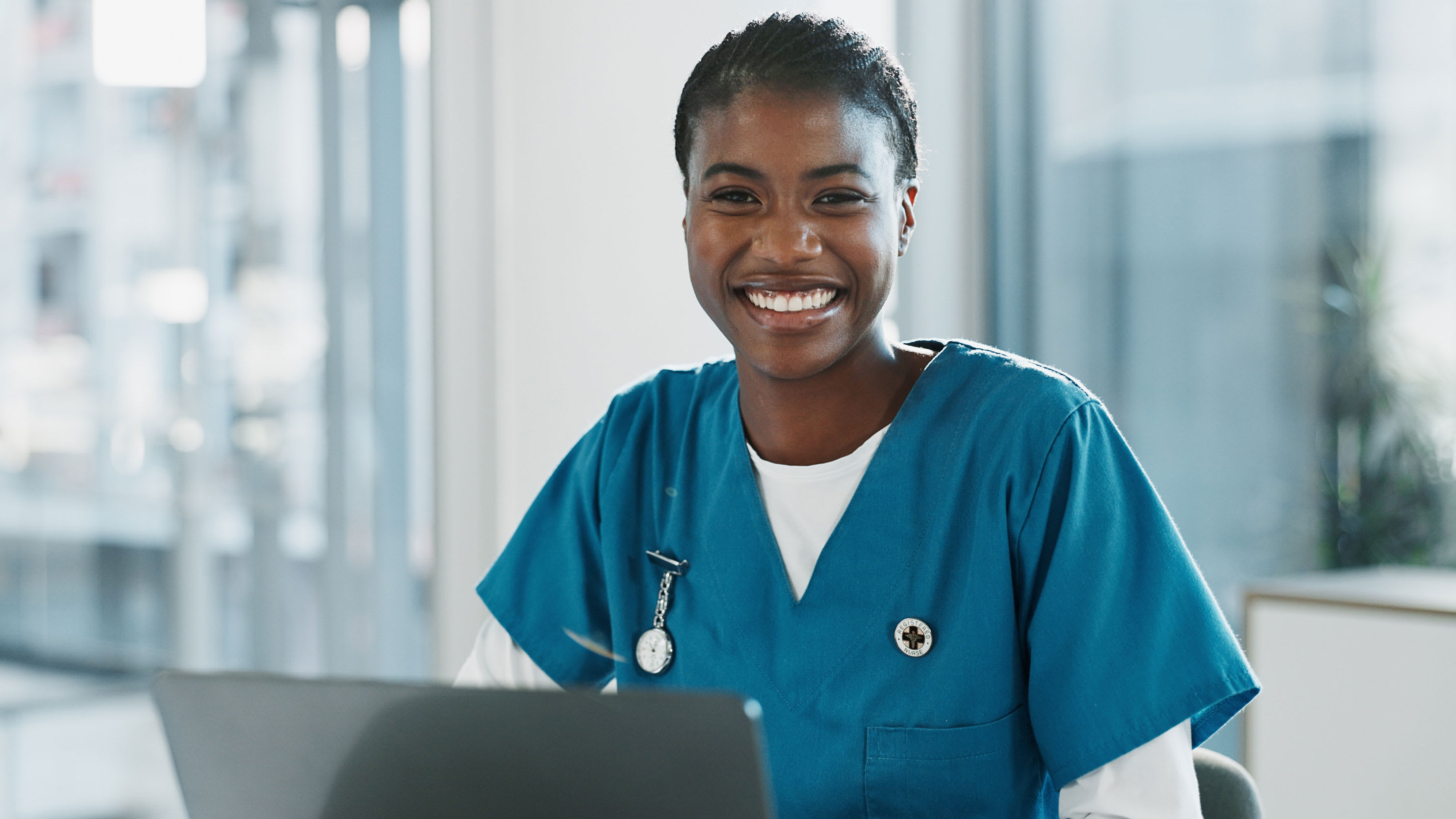 Image of a female physician in scrubs smiling while working at a laptop.