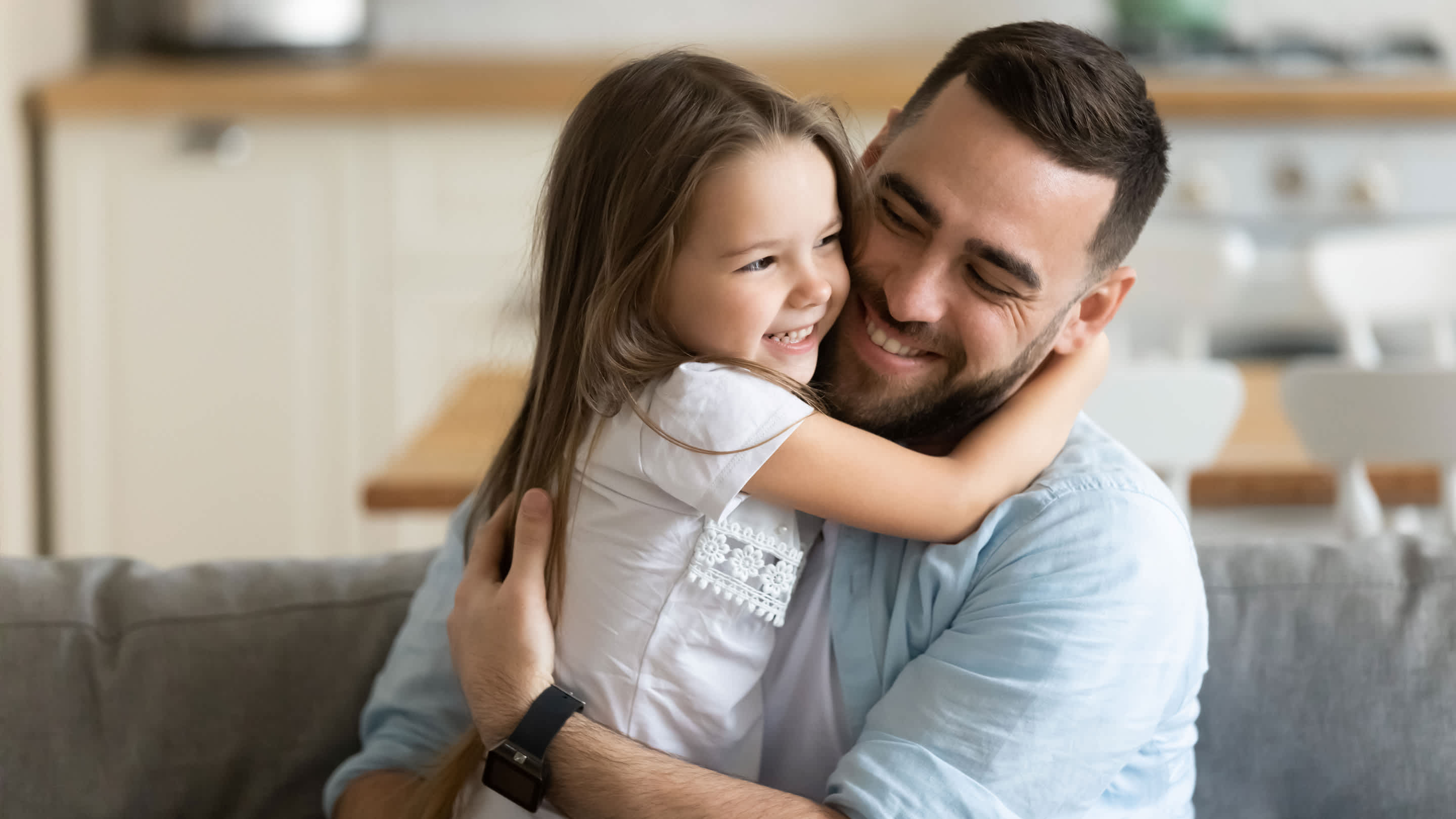 Close up smiling loving young father hugging adorable little daughter, enjoying tender moment, spending weekend together, sitting on cozy couch at home.
