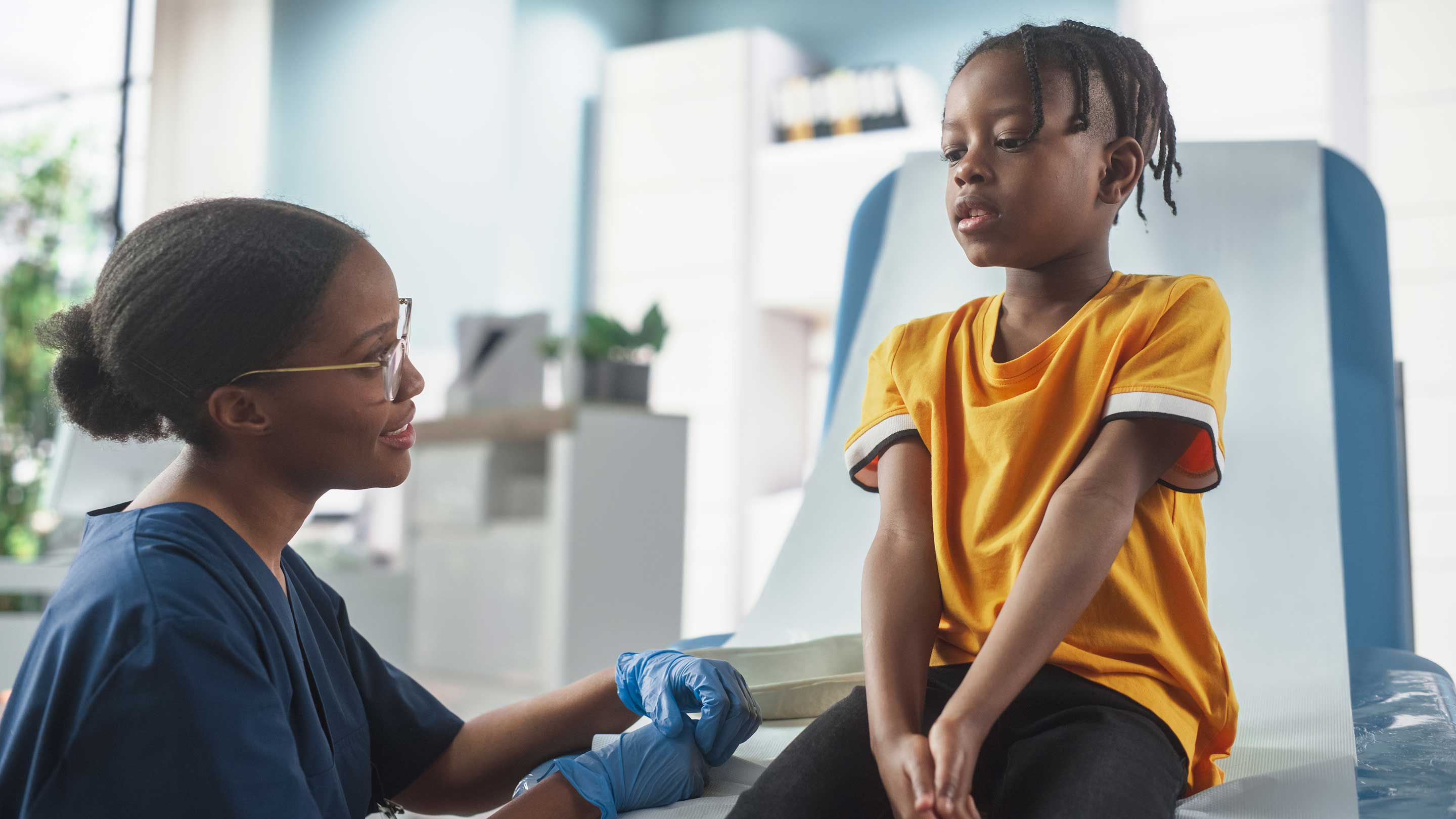 African Boy Sitting In The Chair In Hospital, Talking To Female Nurse.