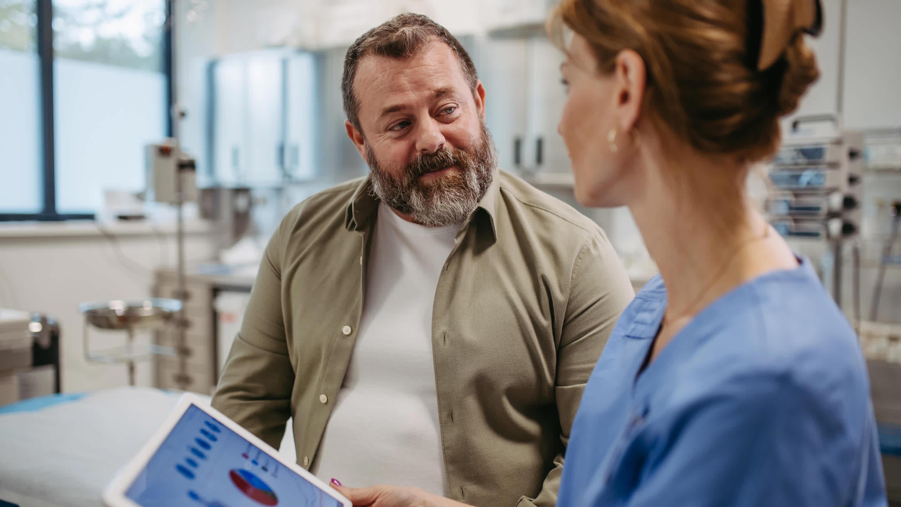 A female doctor stands bedside and talks to an overweight male patient whose back is to the camera.