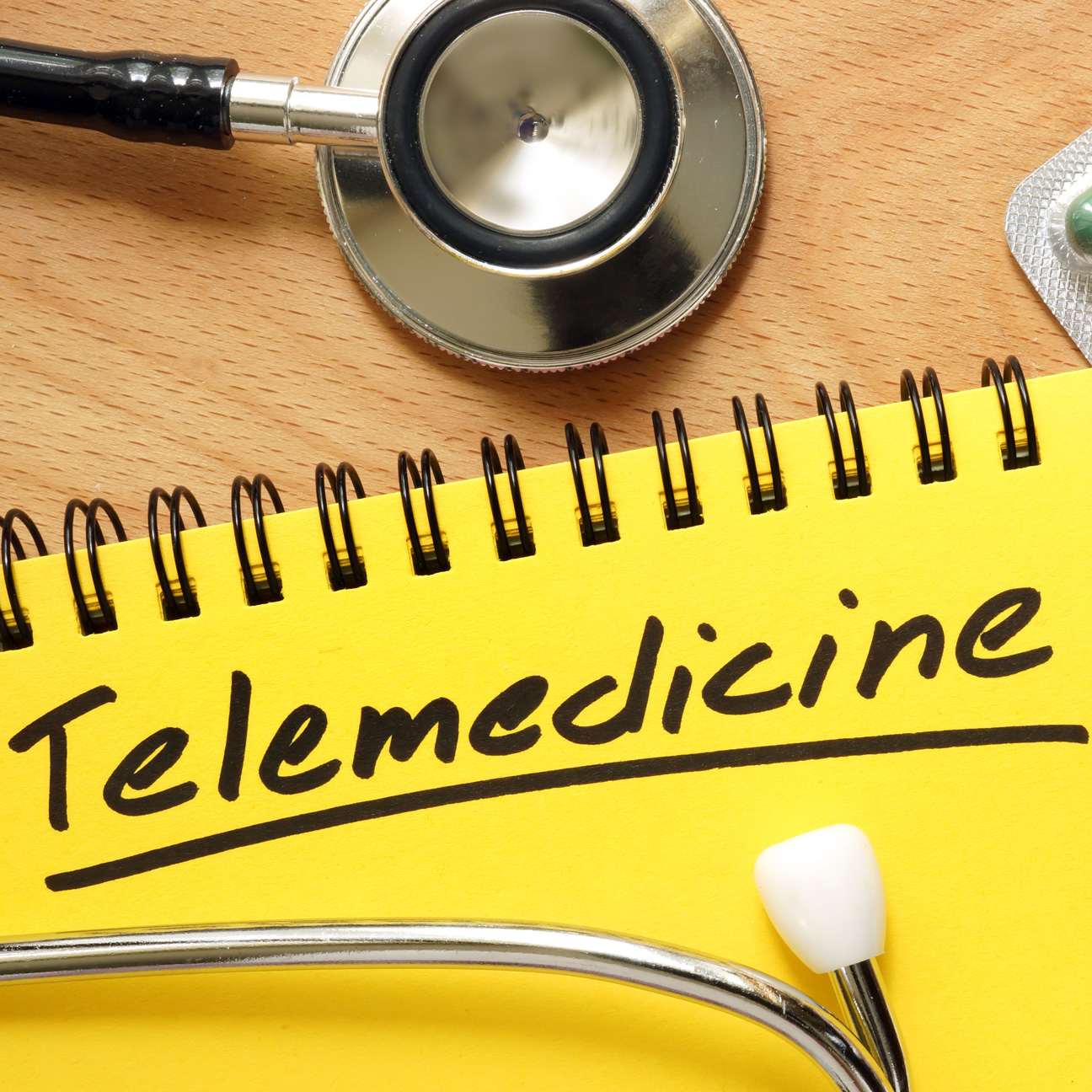 Stethoscope and medicine on a desk with a notebook that reads, “telemedicine”