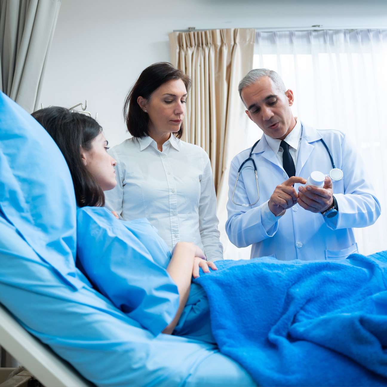 Hospitalist—Physician leading a care team during bedside rounds with a patient in a hospital room.