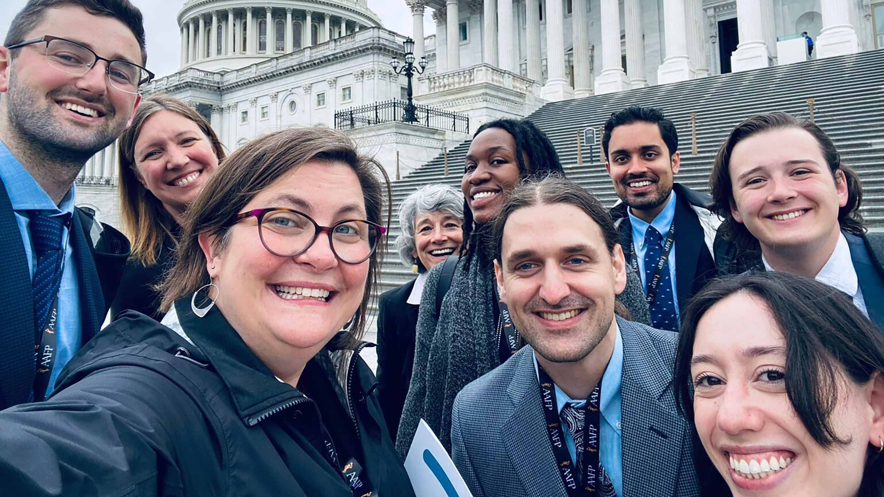 A selfie-style group photo of 9 people in front of the United States Capital Building.