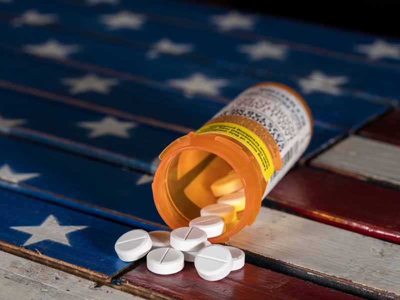 tablets spilling out of pill bottle atop US flag-decorated wooden surface