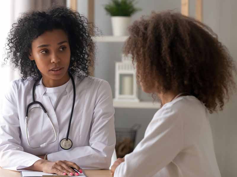 Serious African American female doctor talk to teenage girl sitting at table, focused black woman physician consult teenager, writing down information in patient card. Children health problem concept