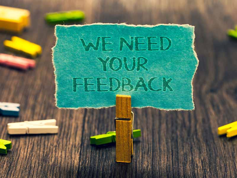 Conceptual hand writing showing We Need Your Feedback. Business photo showcasing Give us your review thoughts comments what to improve Paperclip retain blue cardboard blurry woody desk paper clip