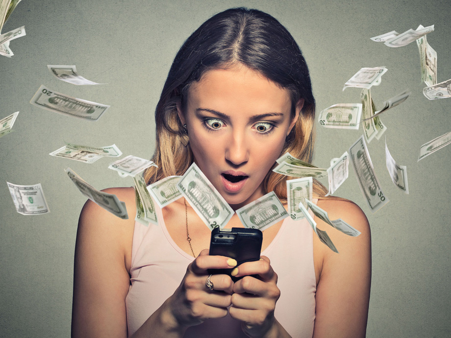 Technology online banking money transfer, e-commerce concept. Shocked young woman using smartphone with dollar bills flying away from screen isolated on gray wall office background.