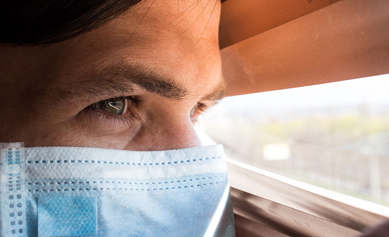 Man privately peering out a window while wearing a surgical mask