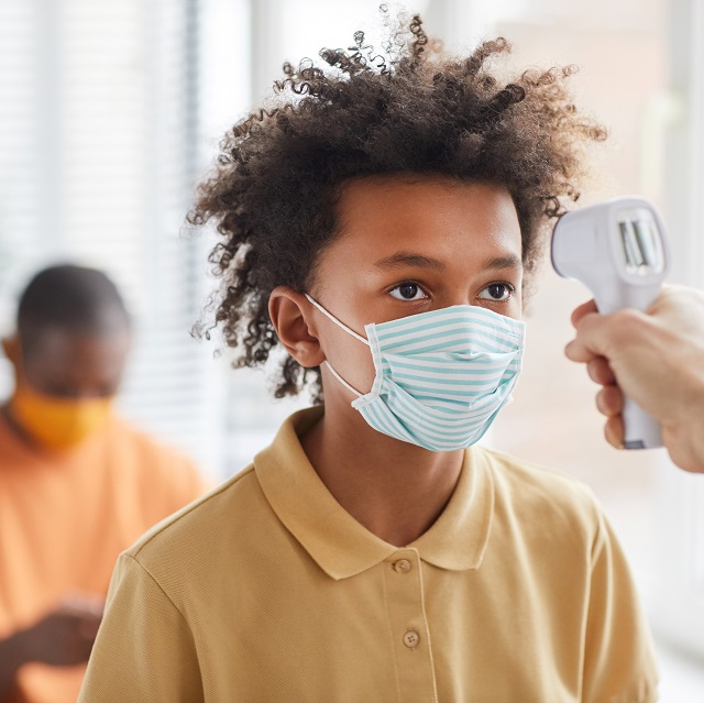 Portrait of teenage African-American boy wearing mask getting temperature check while waiting in line at clinic, copy space
