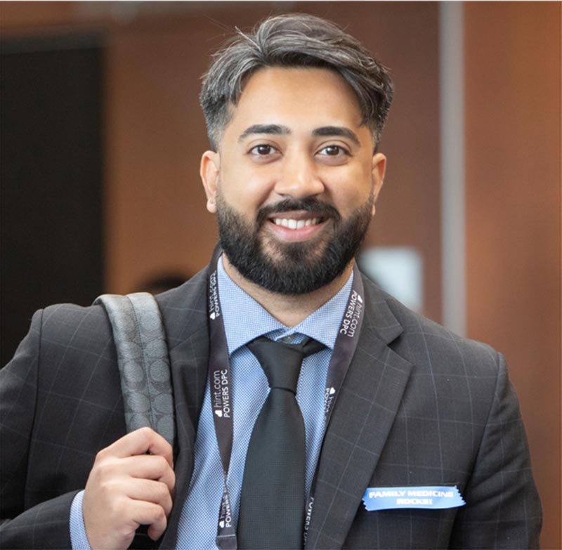 A man with a beard in a suit with a backpack over one shoulder smiling