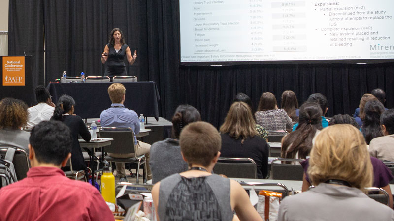 Woman speaking on stage in a convention center room with audience sitting with tables