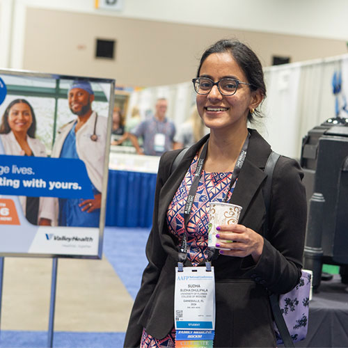An attendee stands smiling with coffee near  standing sign advertising a booth at National Conference 2023.